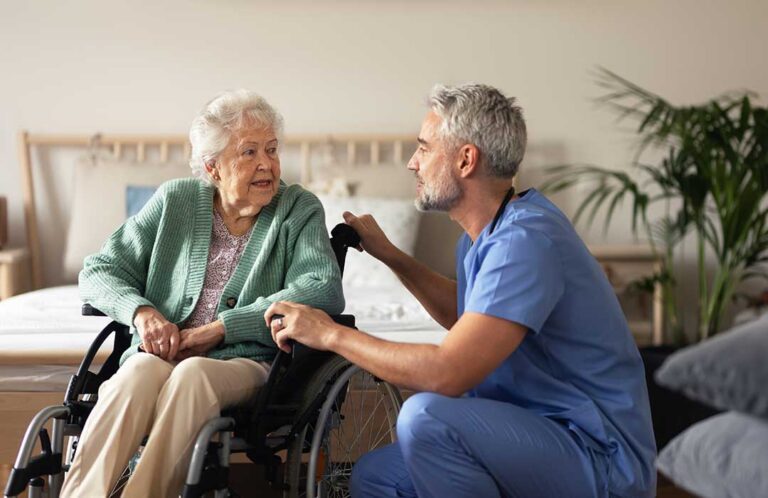 Older adult using a tablet with a caregiver nearby in a bright assisted living room.